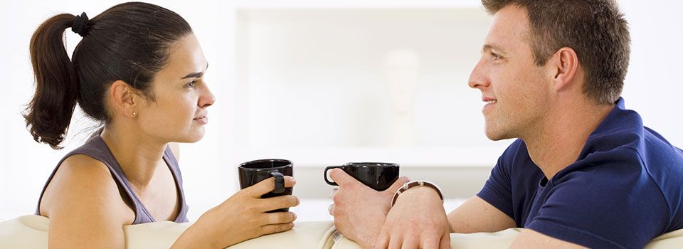 man and woman sitting on couch chatting over a cup of coffee